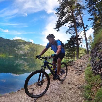 Fietser op mountainbike rijdt langs meerpad bij Crno Jezero in Montenegro, met beboste bergen en blauwe lucht weerspiegeld in water.