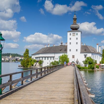 Wit barok kasteel met uivormige koepeltoren op klein eiland, verbonden door houten voetgangersbrug. Groene lantaarnpaal op voorgrond.