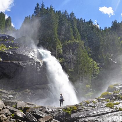 Persoon staat bij de krachtige Krimmler watervallen die over rotsachtige kliffen vallen, omgeven door dicht bos in Oostenrijk.