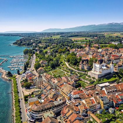 Luchtfoto van Nyon aan de oevers van het Meer van Genève met historisch kasteel, jachthaven, rode pannendaken en Zwitserse Alpen.