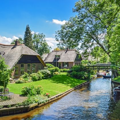 Traditionele rieten huizen met groene tuinen langs een smal kanaal in Giethoorn. Een houten brug kruist de waterweg met boten erbij.