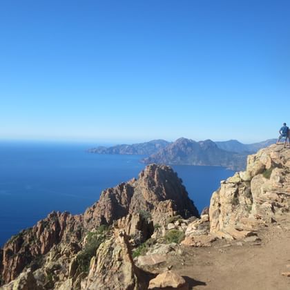 Twee wandelaars op een rotsachtige bergtop in West-Corsica met dramatische rode rotsformaties en blauwe Middellandse Zee op de achtergrond.
