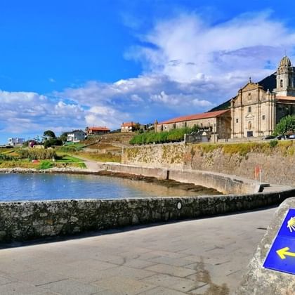 Stenen brug met Camino schelp wegwijzer uitkijkend over Portugees kustdorp. Historische kerk met klokkentoren domineert heuvelvestiging.