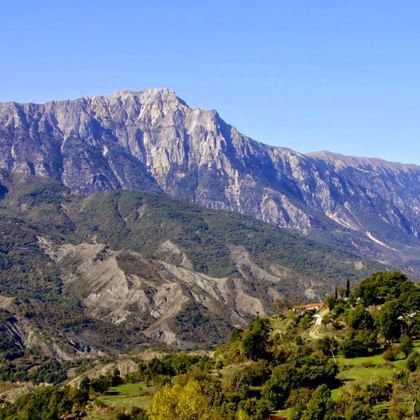 Dramatische rotsachtige bergkam boven groene vallei met verspreide bomen en huizen in Hidden Valley, Albanië, onder heldere hemel.