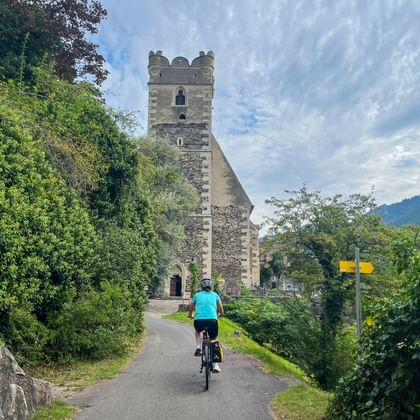 Fietser in turquoise shirt rijdt naar een stenen kerk met toren in Sankt Michael, Wachau. Gele bewegwijzering langs het pad zichtbaar.