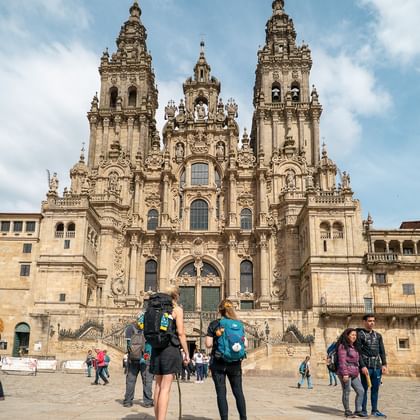 Pelgrims met rugzakken op het plein voor de barokke gevel van de kathedraal van Santiago de Compostela met drie hoge torens onder blauwe lucht.