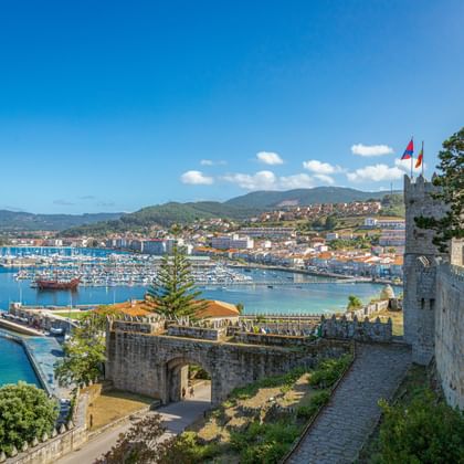 Uitzicht vanaf stenen vestingmuren in Baiona op jachthaven met zeilboten, kustplaats en groene heuvels onder blauwe lucht.