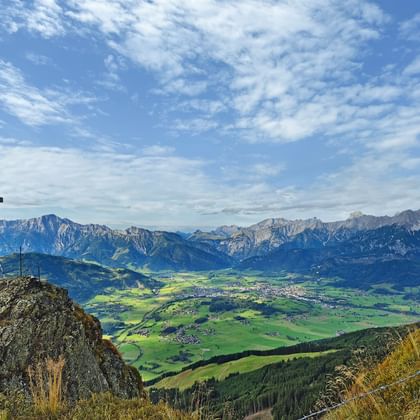 Topkruis op rotsachtige top met uitzicht op groene vallei en Kitzbüheler Alpen onder blauwe lucht met verspreide wolken.