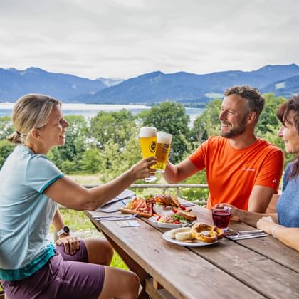 Drie fietsers proosten met bier aan een houten tafel met uitzicht op de Tegernsee. Traditionele Beierse snacks met bergzicht op de achtergrond.
