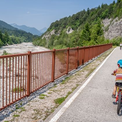 Kind met blauwe helm fietst op Alpe Adria fietspad langs rivier in Kanaltal vallei, met bergen en rode reling zichtbaar.
