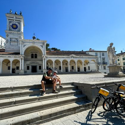 Twee fietsers zitten op trappen op Piazza della Libertà, Udine, met fietsen en gele tassen in de buurt. Historische klokkentoren en arcaden zichtbaar.