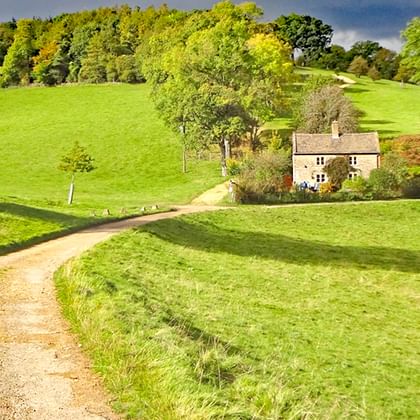 Stenen cottage genesteld in glooiende groene heuvels van de Cotswolds. Een onverharde weg leidt door weilanden naar het huis, omringd door bomen.
