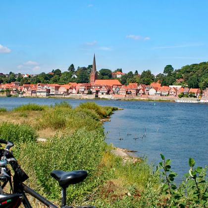 Zwarte fiets met tassen geparkeerd aan rivieroever met uitzicht op Lauenburg aan de Elbe, met historische gebouwen en kerktoren.