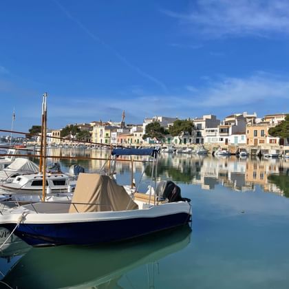 Rustige haven van Cala Ratjada met vissersboten en jachten afgemeerd in turquoise water, kleurrijke mediterrane gebouwen weerspiegeld.