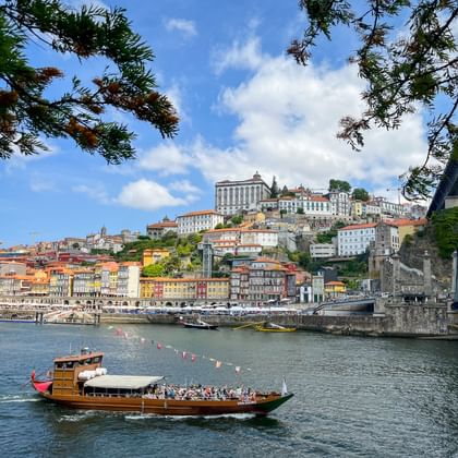 Traditionele houten boot op de Douro met Porto's kleurrijke gebouwen op de heuvel en brug. Blauwe lucht met witte wolken boven het stadsgezicht.