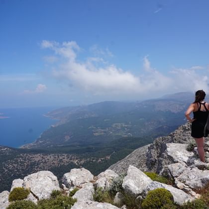 Wandelaar op rotsachtige bergtop op Rhodos met uitzicht op blauwe Egeïsche Zee, groene valleien en verre kustlijn onder heldere hemel.