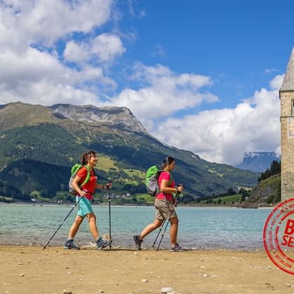 Wandelaars bij Reschensee met klokkentoren Twee wandelaars met rugzakken en wandelstokken steken ondiep water over bij de beroemde klokkentoren van Graun in het Reschensee.
