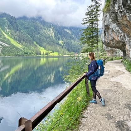 Vrouwelijke wandelaar met blauwe rugzak op verharde oeverpad bij Gosauseen, met houten leuning, bergmeer en beboste bergen.