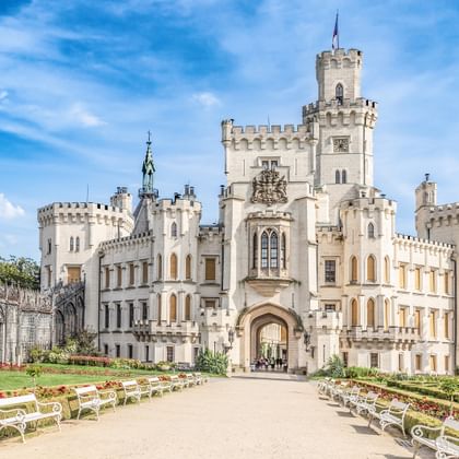 Wit neogotisch kasteel Hluboka met torens en kantelen, omgeven door formele tuinen met bloemperken en witte banken onder blauwe hemel.