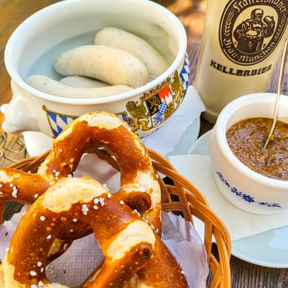 Traditioneel Beiers ontbijt met witte worsten in decoratieve kom, zoutpretzel, zoete mosterd en Kellerbier-fles op houten tafel.