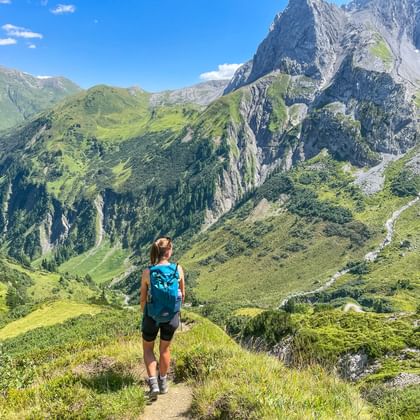 Wandelaar met blauwe rugzak op bergpad in de Allgäu Alpen, uitkijkend over groene valleien en dramatische rotspieken onder blauwe lucht.