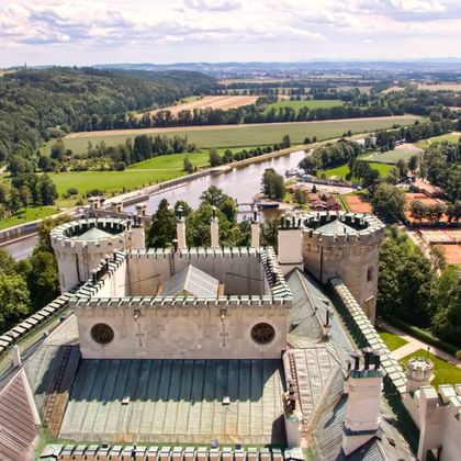 Luchtfoto van wit kasteel met kantelen en turquoise dak in Teplice nad Bečvou, met uitzicht op groene velden, rivier en beboste heuvels.
