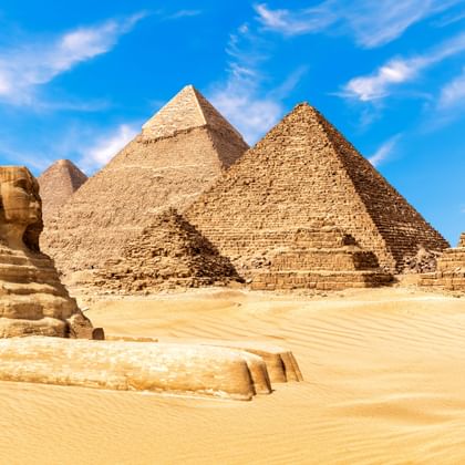 De Grote Sfinx op de voorgrond met drie piramides van Gizeh die oprijzen uit gouden woestijnzand onder een blauwe lucht met witte wolken in Egypte.
