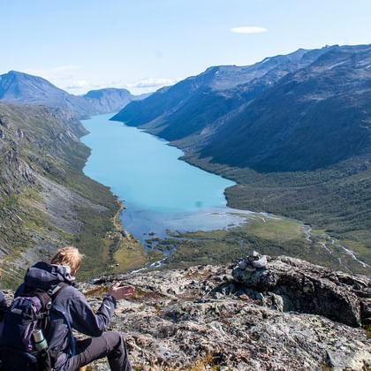 Drie wandelaars met rugzakken zitten op rotsachtige top en kijken uit over turquoise meer omringd door bergen in Jotunheimen, Noorwegen.