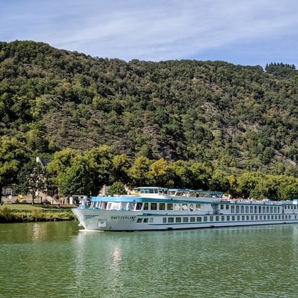 MS Switzerland riviercruiseschip varend op kalm groen water met beboste heuvels op de achtergrond onder een gedeeltelijk bewolkte hemel.