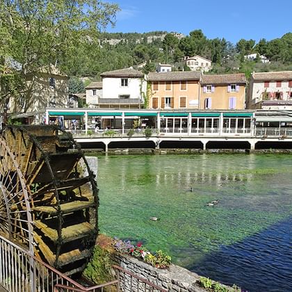 Historisch houten watermolen naast de heldere rivier Sorgue in L'Isle-sur-la-Sorgue, met kleurrijke Provençaalse huizen en cafés langs het water.