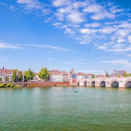 Meerbogige stenen brug over de Maas met historische gebouwen en kleurrijke huizen langs de waterkant onder blauwe hemel.