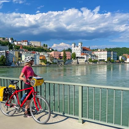 Fietser met rode fiets en gele fietstassen op brug met uitzicht op Passau langs de Donau onder blauwe hemel met wolken.