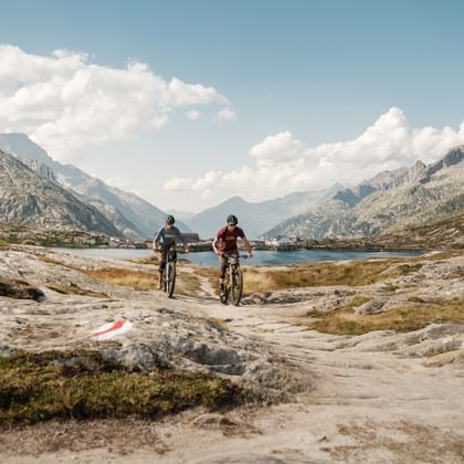Twee mountainbikers rijden op een rotsachtig pad in de Zwitserse Alpen met een bergmeer en dramatische toppen op de achtergrond onder bewolkte luchten.