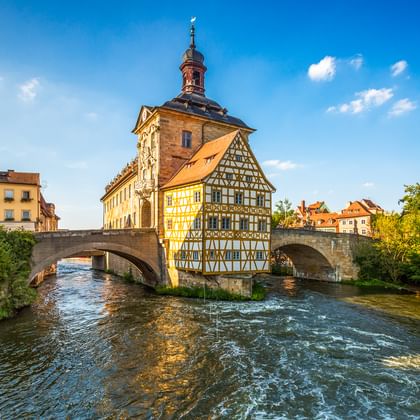 Historisch vakwerkhuis van het Oude Stadhuis van Bamberg op eiland in de Regnitz, verbonden door stenen brugbogen onder blauwe lucht.