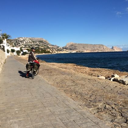 Fietser op kustpromenade in Denia Fietser met toerfiets op stenen promenade langs Middellandse Zeekust in Denia, met blauwe zee, rotsachtige kust en witte gebouwen.