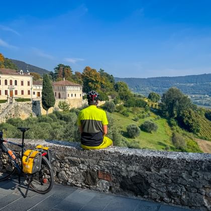 Fietser in geel shirt zit op stenen muur met uitzicht op Italiaans landschap. Toerfiets met gele tassen staat ernaast. Historische gebouwen en heuvels zichtbaar.