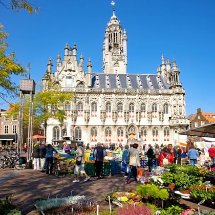 Gotisch stadhuis van Middelburg met sierlijke torens en decoratieve gevel. Marktkramen met bloemen en winkelende mensen op het plein.