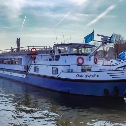 Wit-blauw riviercruiseschip MS Clair d'Etoile afgemeerd in een haven met andere boten zichtbaar op de achtergrond onder een bewolkte hemel.