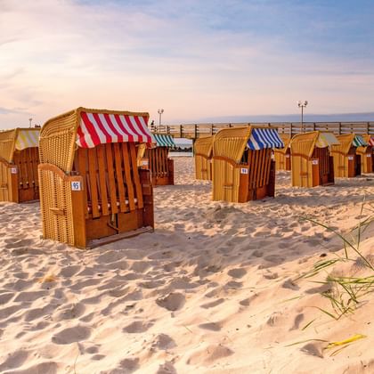 Rij traditionele rieten strandkorfjes met gestreepte kappen op zandstrand in Wustrow. Pier zichtbaar op achtergrond onder avondlucht.