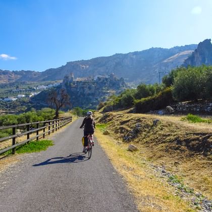 Fietser op geasfalteerd spoorwegpad in Andalusië met houten leuningen, bergen en historische bergstad zichtbaar op achtergrond onder blauwe lucht.