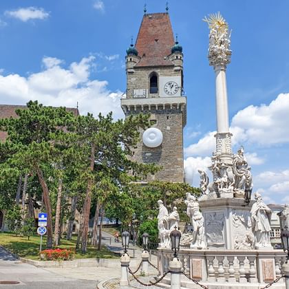 Kasteel Perchtoldsdorf met middeleeuwse stenen toren en klok, naast een sierlijke barokke pestkolom met witte sculpturen op een stadsplein.
