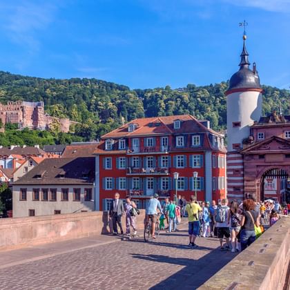 Toeristen wandelen over Heidelbergs Oude Brug met tweeling torens en poort. Kasteel Heidelberg zichtbaar op beboste heuvel.