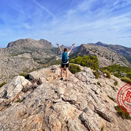 Persoon met opgeheven armen staat op rotsachtige bergtop met uitzicht op dramatisch berglandschap onder blauwe hemel.