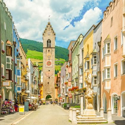 Kleurrijke historische gebouwen langs een voetgangersstraat in Sterzing, Italië, met een hoge middeleeuwse toren in het midden en groene bergen op de achtergrond.