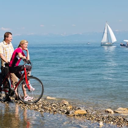 Twee fietsers met beladen fiets op rotsachtige oever van het Bodenmeer. Zeilboot en motorboot zichtbaar op blauw water met bergen op achtergrond.
