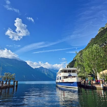 Wit-blauwe passagiersveerboot aangemeerd in Riva del Garda aan het Gardameer, met bergen en kleurrijke gebouwen langs de kust onder blauwe hemel.