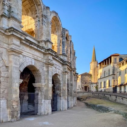 Oud Romeins amfitheater in Arles met stenen bogen en zuilen. Historische gebouwen en kerktoren zichtbaar op de achtergrond onder blauwe lucht.