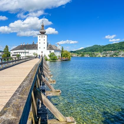 Houten loopbrug naar kasteel Schloss Orth aan de Traunsee in Salzkammergut. Helder blauw water, wit kasteel met toren, groene heuvels.