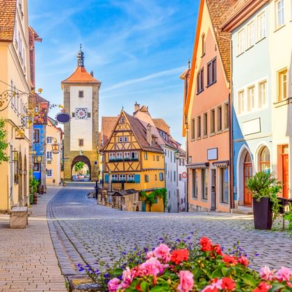 Geplaveide straat in Rothenburg met kleurrijke vakwerkhuizen en de witte Siebersturm toren met oranje dak onder blauwe hemel.