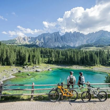 Twee fietsers met rugzakken staan bij een houten hek met uitzicht op een turquoise bergmeer in de Dolomieten, met dicht bos en rotspieken.